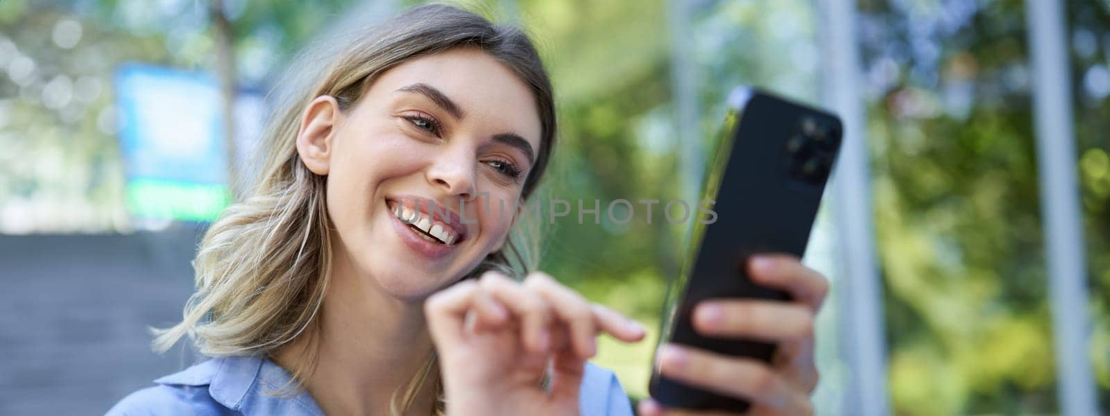 Beautiful young woman sitting outdoors on street, smiling at smartphone, buying online, order on mobile phone.