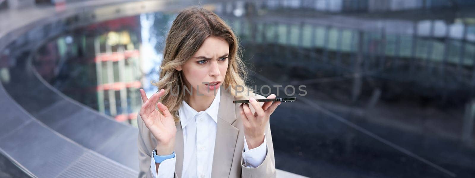 Woman looks angry while leaving voice message on mobile phone, talks into speakerphone with frowned face.