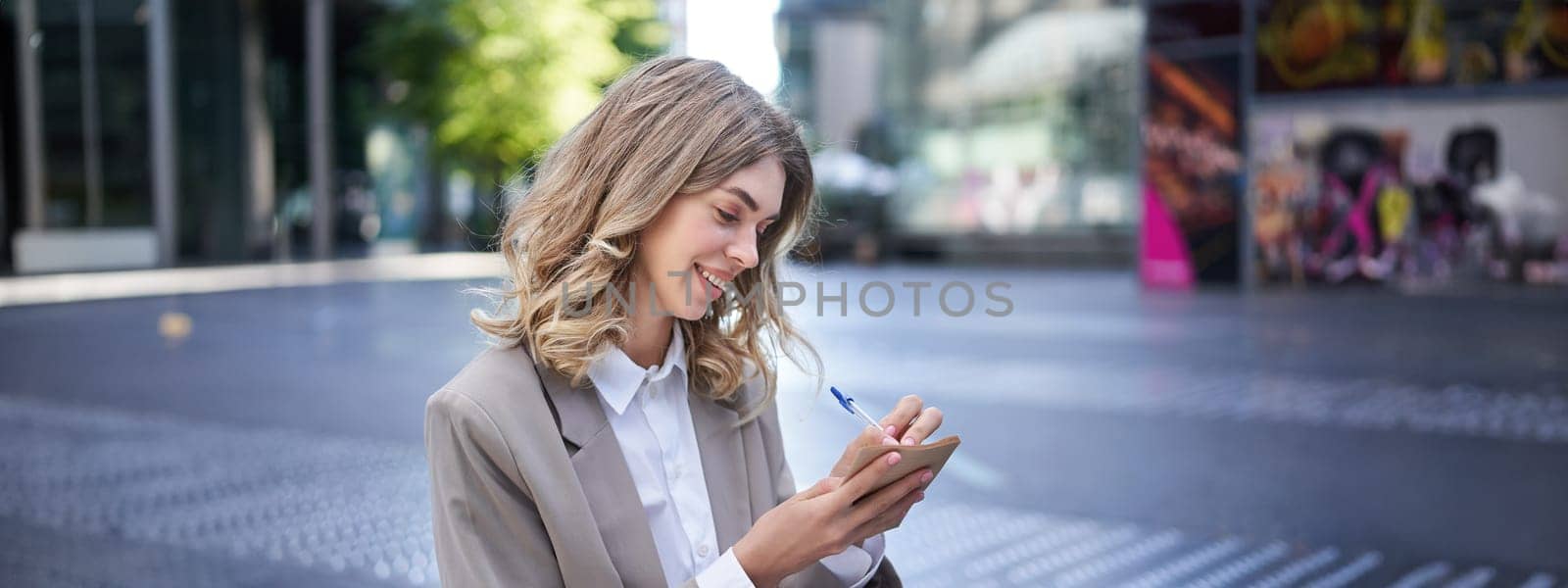 Young businesswoman preparing for speech, interview in company, writing down notes while sitting outside in city centre by Benzoix