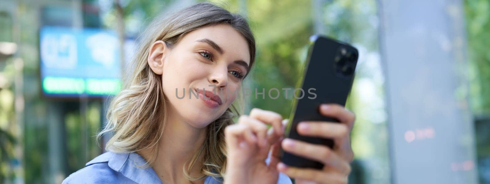 Female employee sitting outdoors and looking at mobile phone, smiling at smartphone screen.