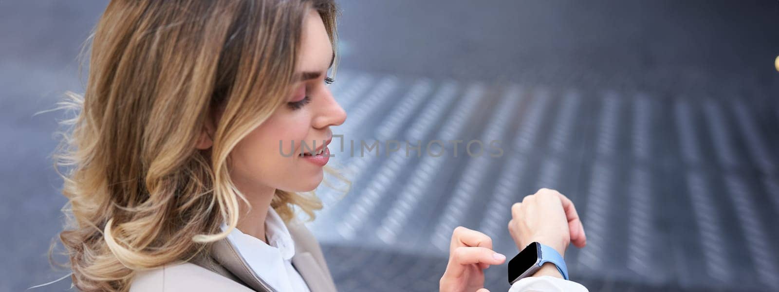 Close up of businesswoman reading message on digital watch. Office lady in suit checking time and smiling, standing on street.