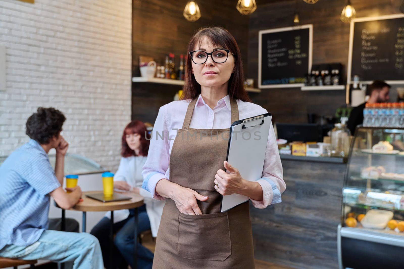 Portrait of confident middle aged woman coffee shop owner. Female in apron holding clipboard with orders looking at camera inside cafe hall, resting clients working barista. Small business work people