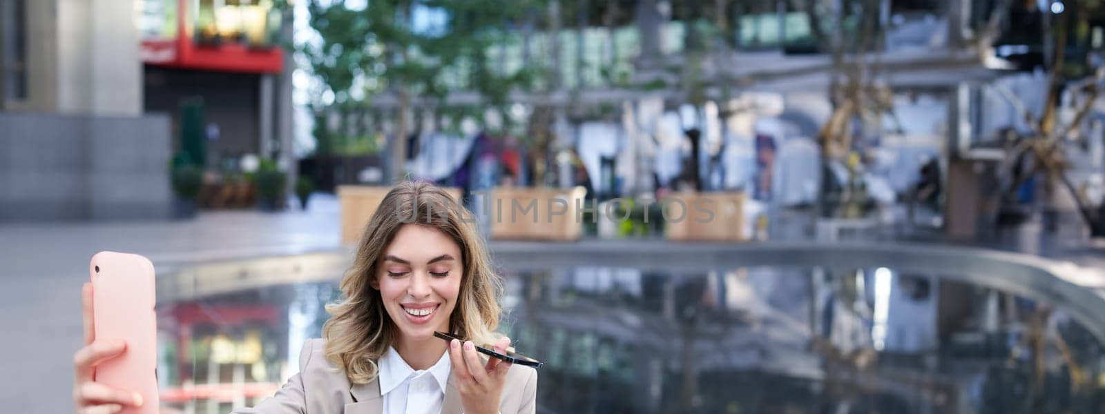 Vertical shot of smiling woman records voice message and looks at digital watch, reads message or checks time, sits in city centre by Benzoix