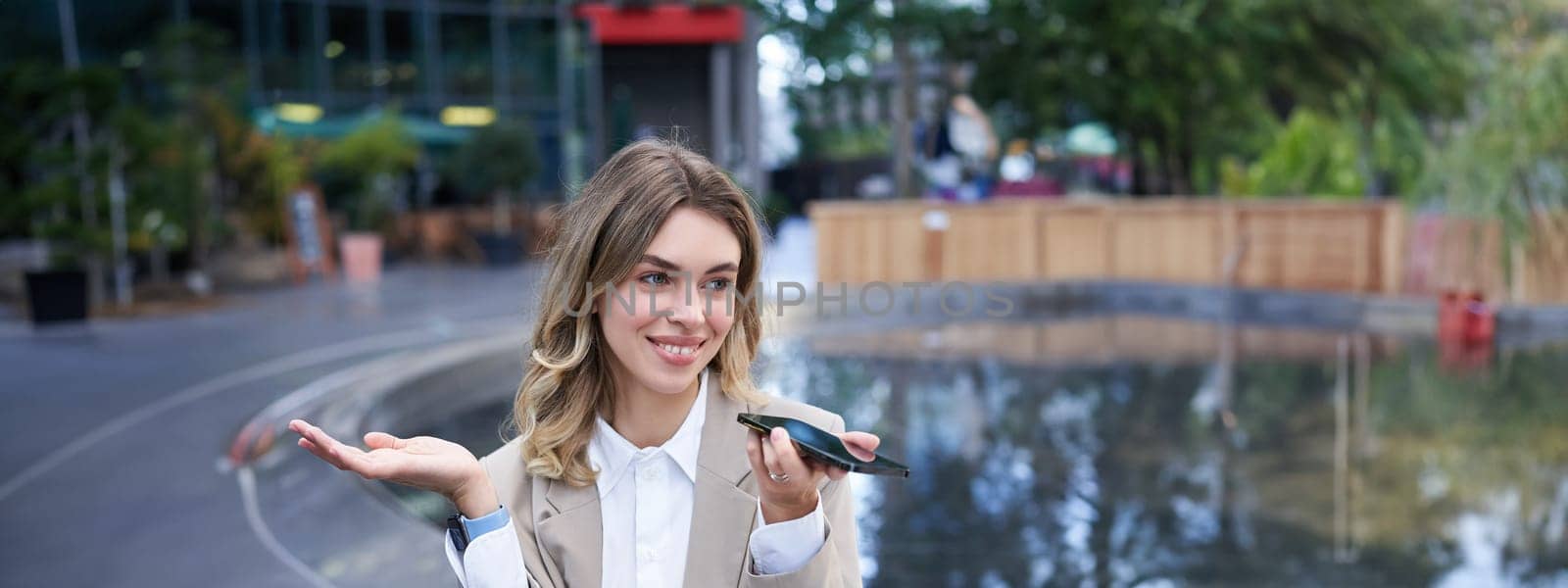 Smiling business woman record voice message, speaking into microphone on mobile phone, sitting near fountain on street by Benzoix