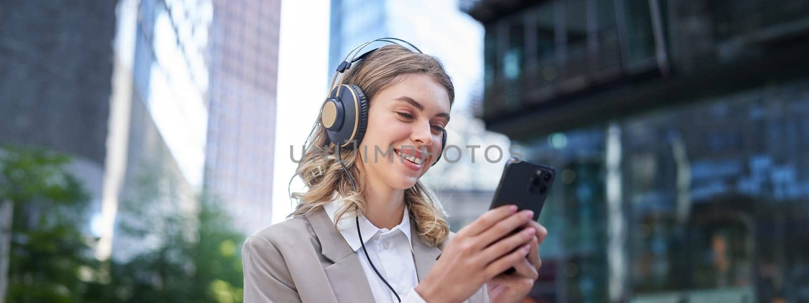 Portrait of smiling young businesswoman listening music in headphones and using mobile phone while on a busy city street.