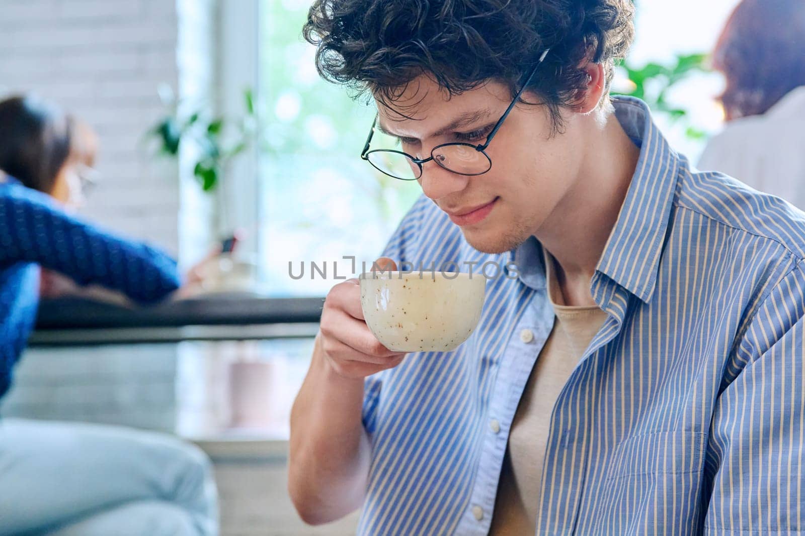 Handsome young curly-haired guy drinking cup of coffee in coffee shop. Young male college student in glasses enjoying cappuccino. Coffee culture, lifestyle, youth, people concept