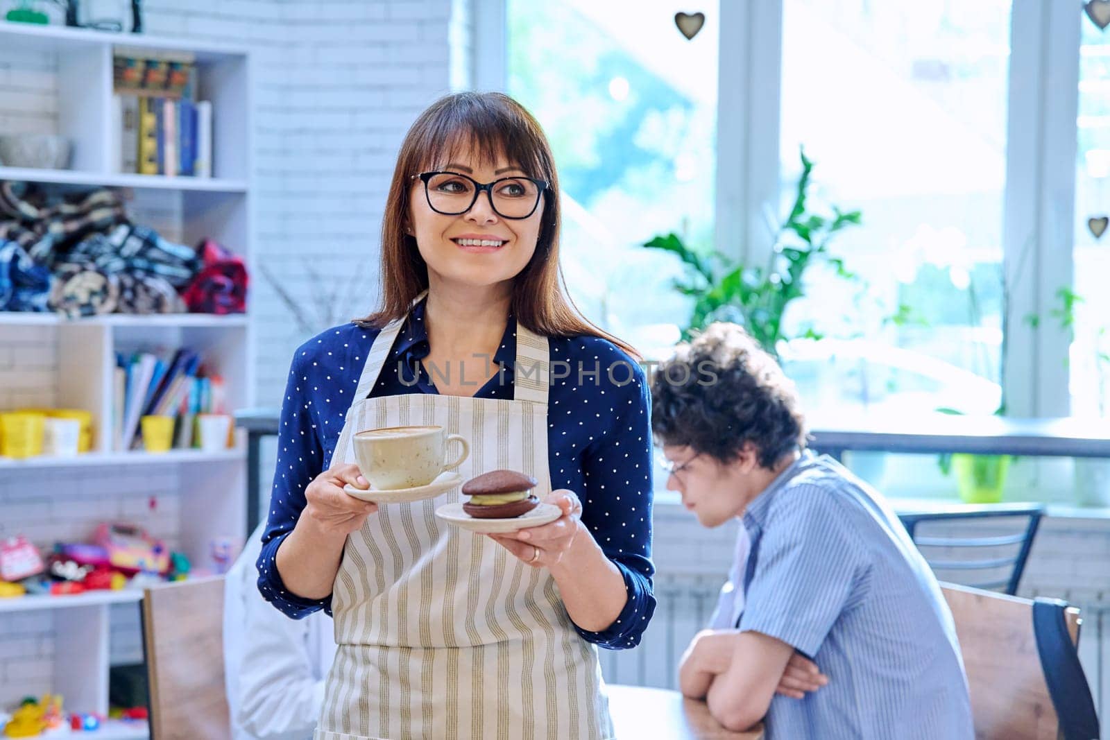 Portrait of female in apron holding fresh cup of coffee and cake on plate in coffee shop by VH-studio