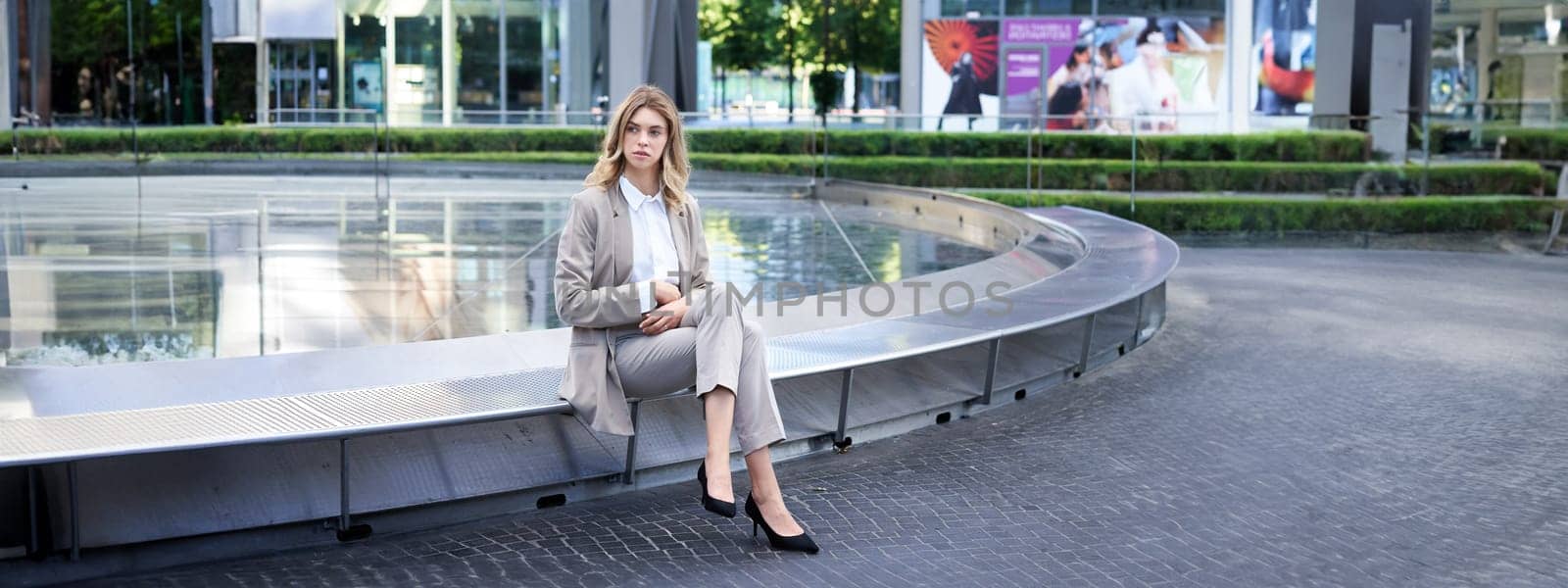 Businesswoman sitting alone on empty street near fountain. Female entrepreneur outdoors in suit and heels.