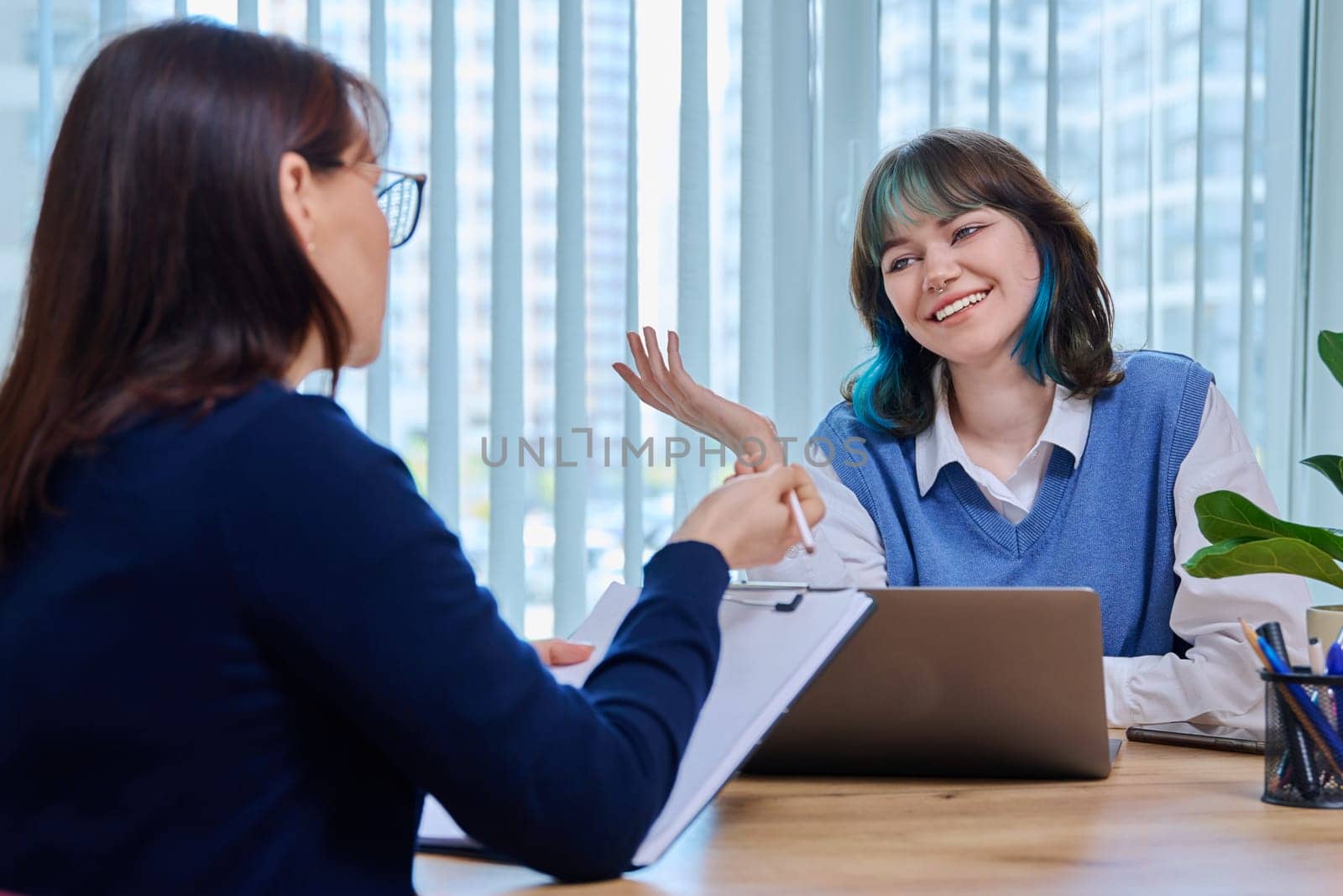 Middle aged woman teacher mentor talking helping testing female teenage student sitting in office in college building. Education, learning, contact teacher student concept