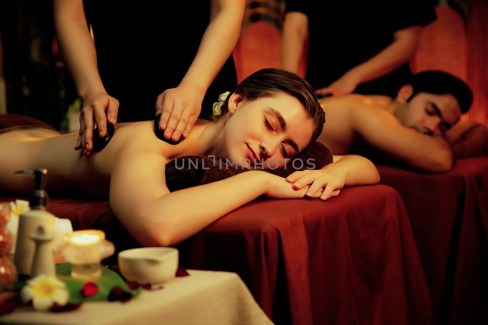 Hot stone massage at spa salon in luxury resort with warm candle light, blissful couple customer enjoying spa basalt stone massage glide over body with soothing warmth. Quiescent