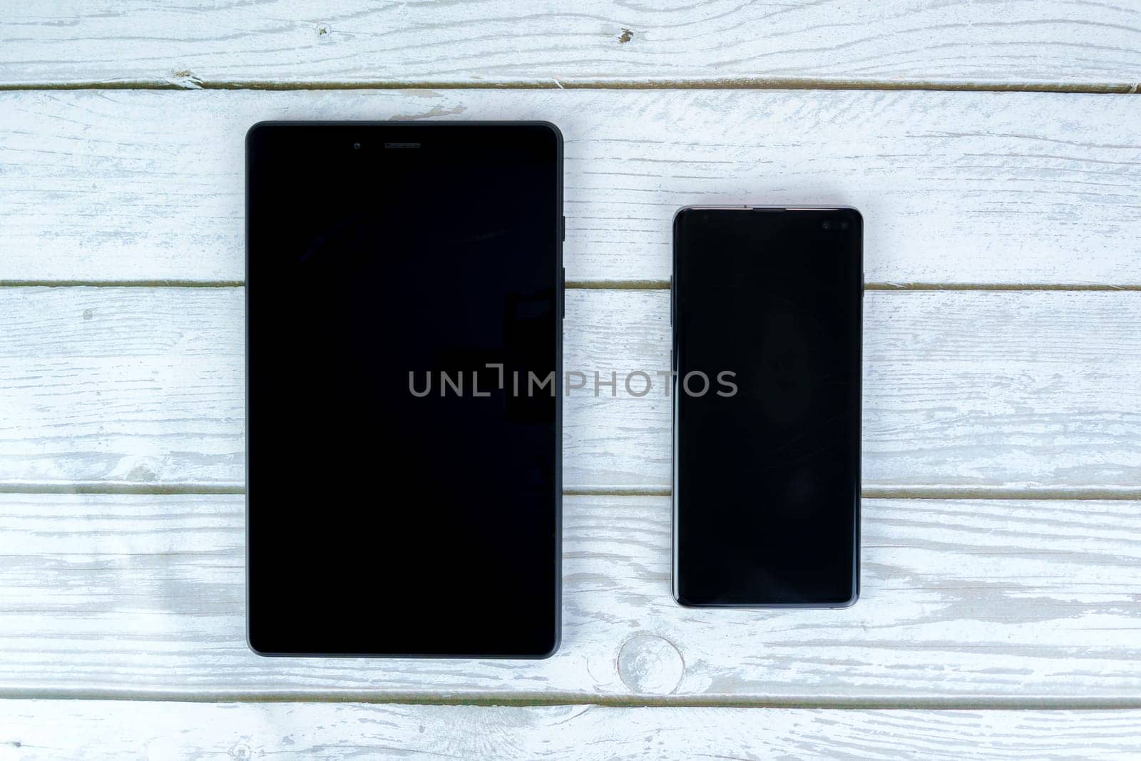 Black tablet and smartphone with turned-off screens placed side by side on a white wooden surface. Top-down view.