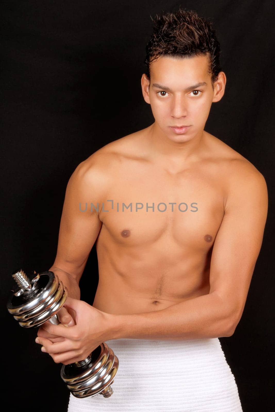Shirtless young man training with a chrome dumbbell over a black background. Soft focus.