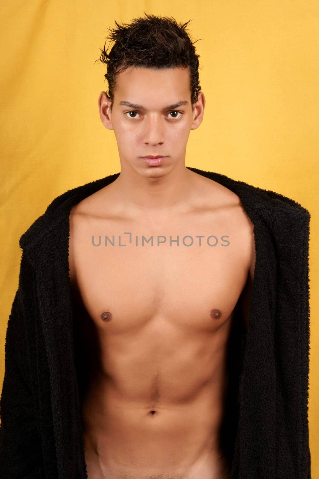 Sexy brazilian young man with black bathrobe over yellow background.
