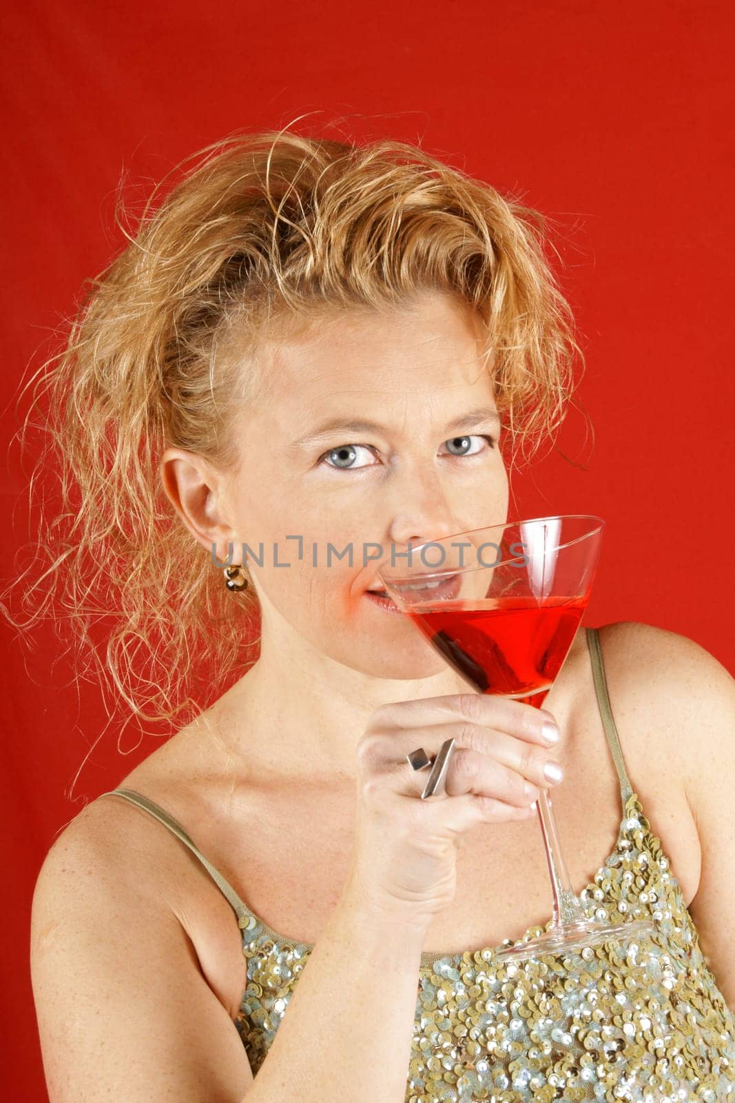 Portrait of a beautiful blond woman with blue eyes, wearing a green sequins top, holding a red drink, over a red background. Soft focus.