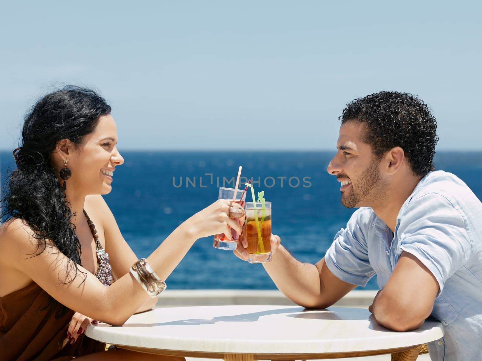 honeymoon: young just married hispanic couple sitting at bar table near the sea and drinking cocktail. Horizontal shape, side view, copy space
