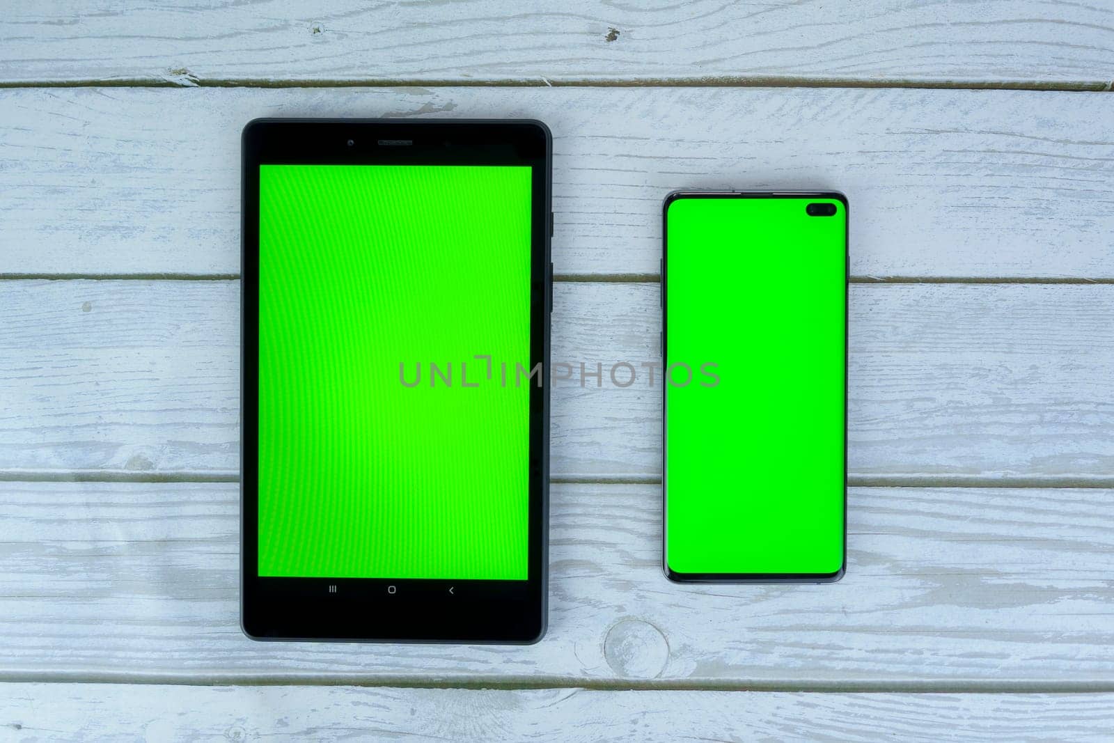 Black tablet and smartphone with blank green screens lying on a white wooden surface, ready for mockup or design presentation.