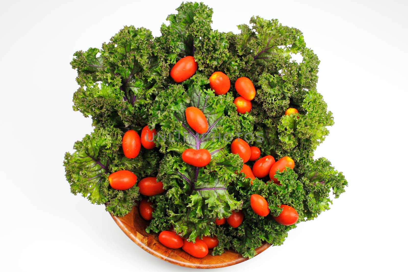 Small red cherry tomatoes on top of red kale in a wood bowl on white background.