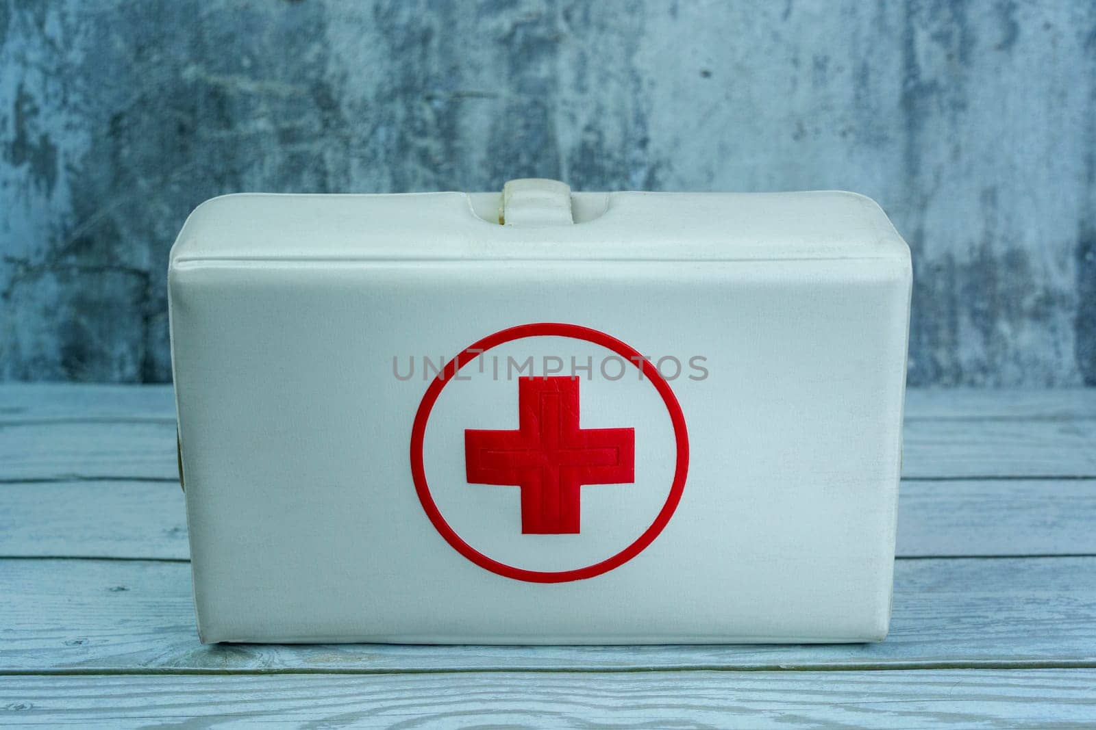 Vintage white first aid kit box with a red cross symbol, placed on a wooden surface against a gray textured background. Ideal for medical and emergency themes.