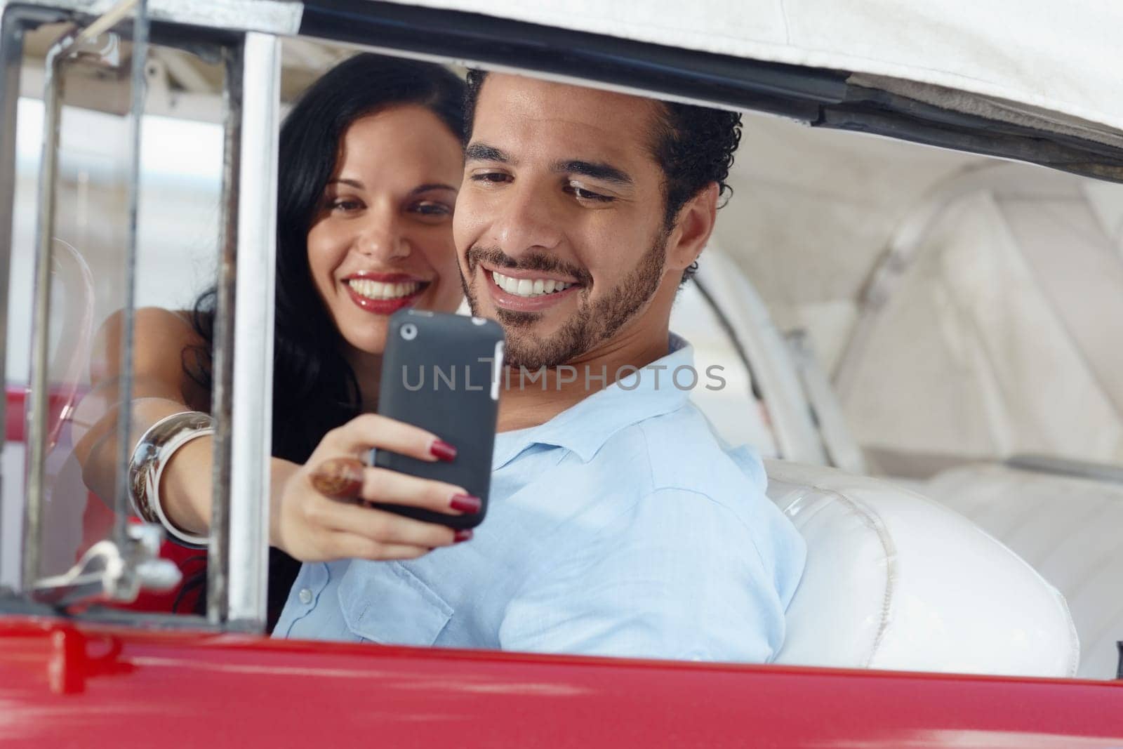 happy young man and woman smiling while taking snapshot with cell phone camera from red vintage convertible car