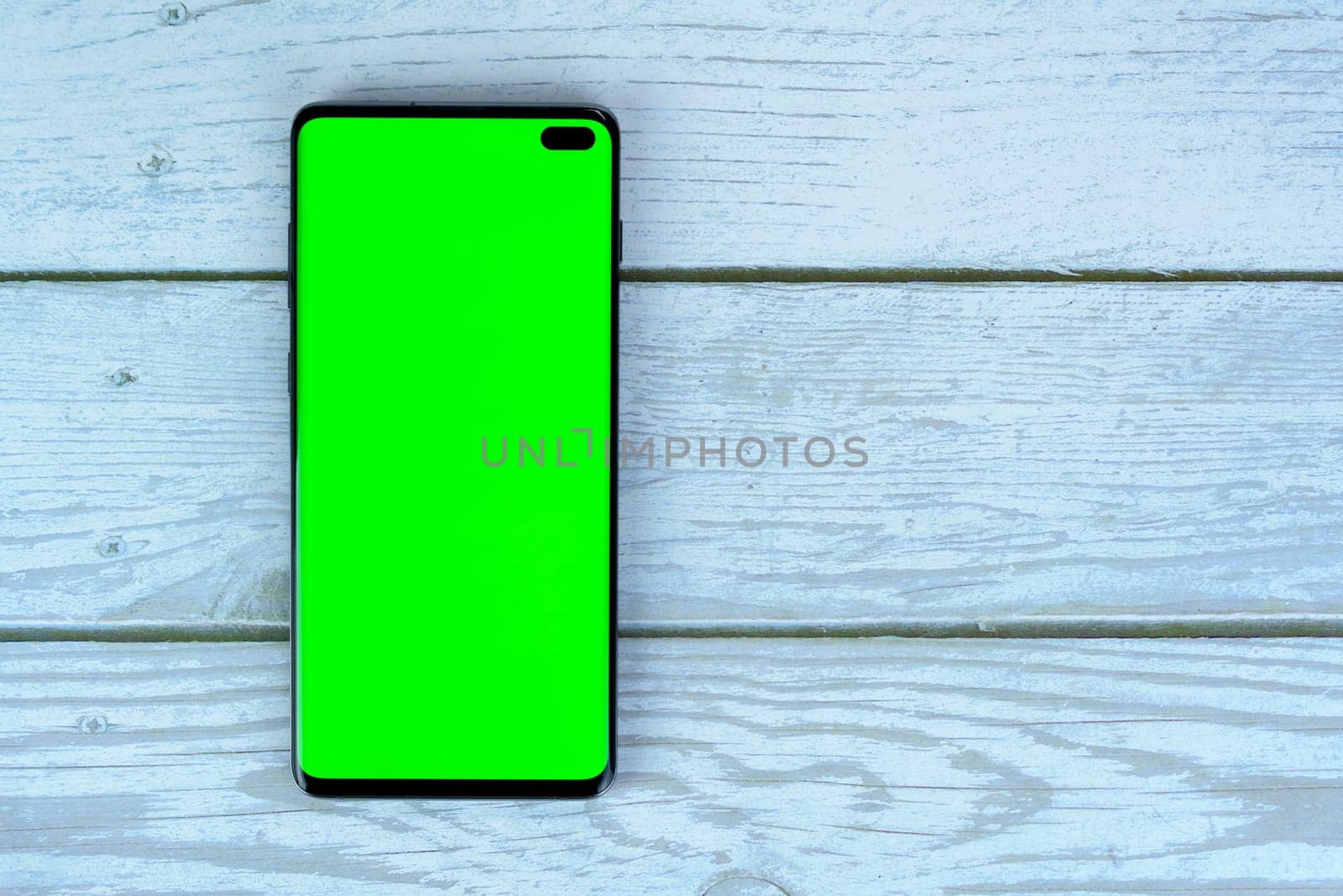 Black smartphone with blank green screen lying on a white wooden surface, ideal for mockups, design presentations, or advertising purposes.