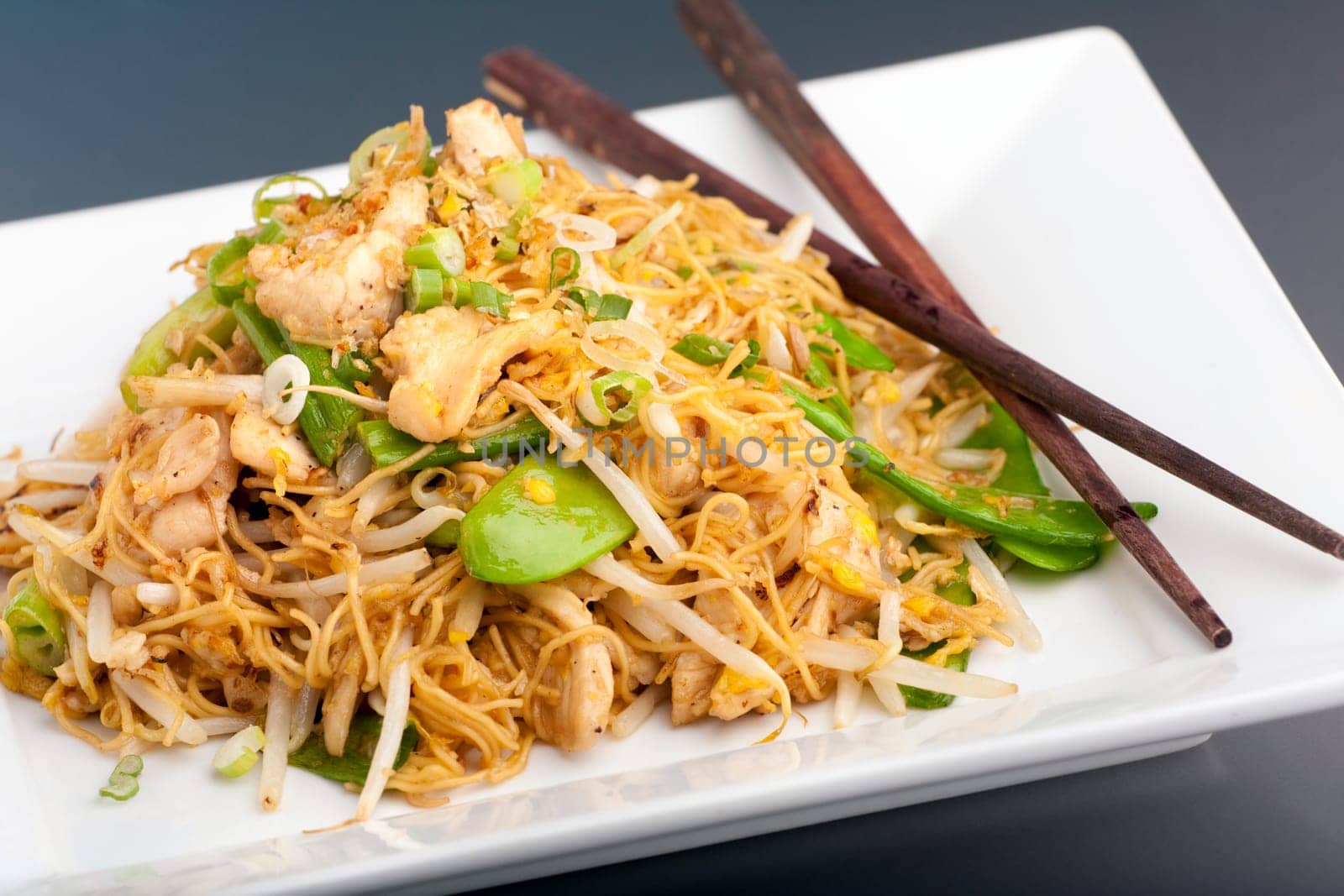 A Thai dish of chicken and noodles stir fry presented on a square white plate with wooden chopsticks.