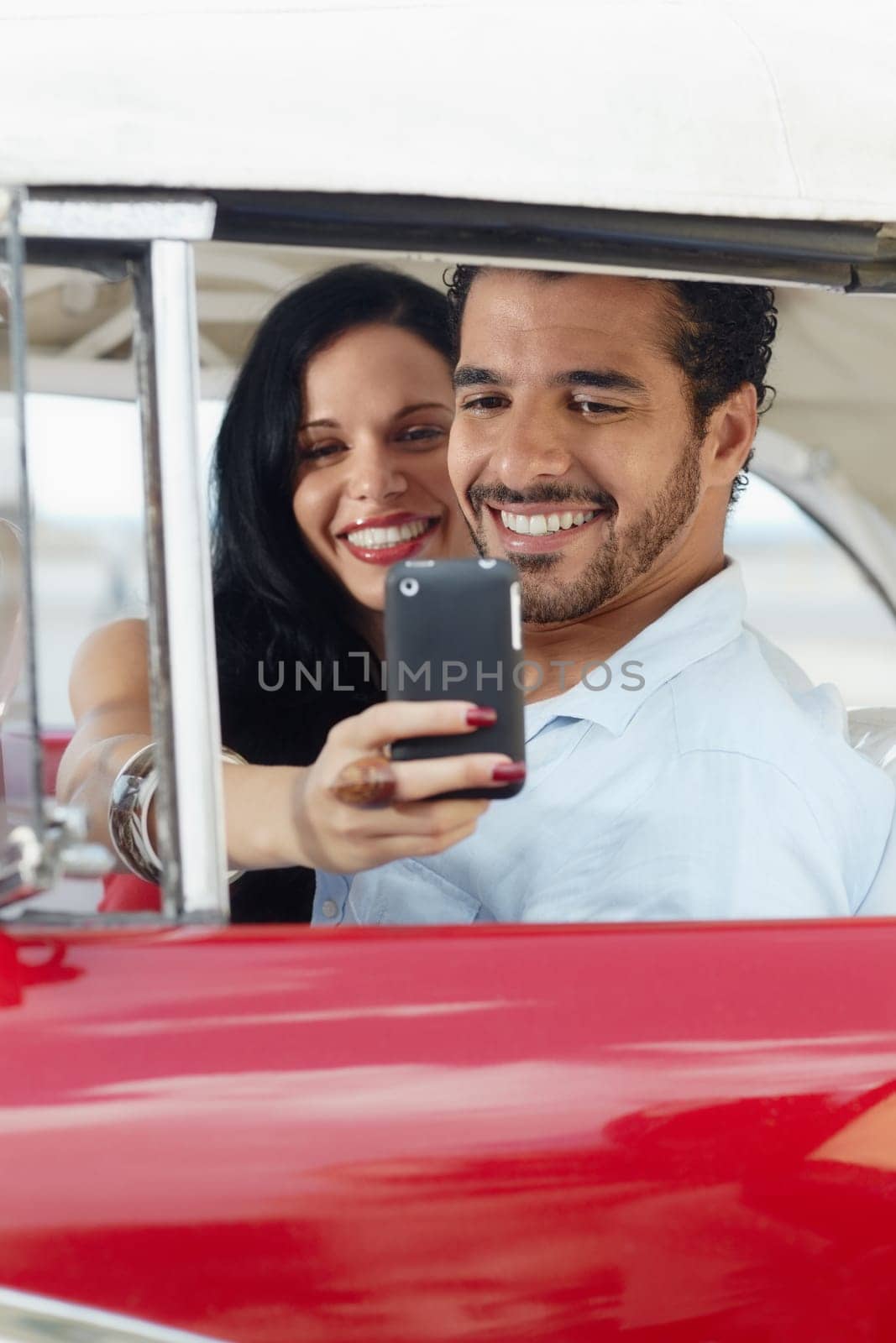 happy young man and woman smiling while taking snapshot with cell phone camera from red vintage convertible car