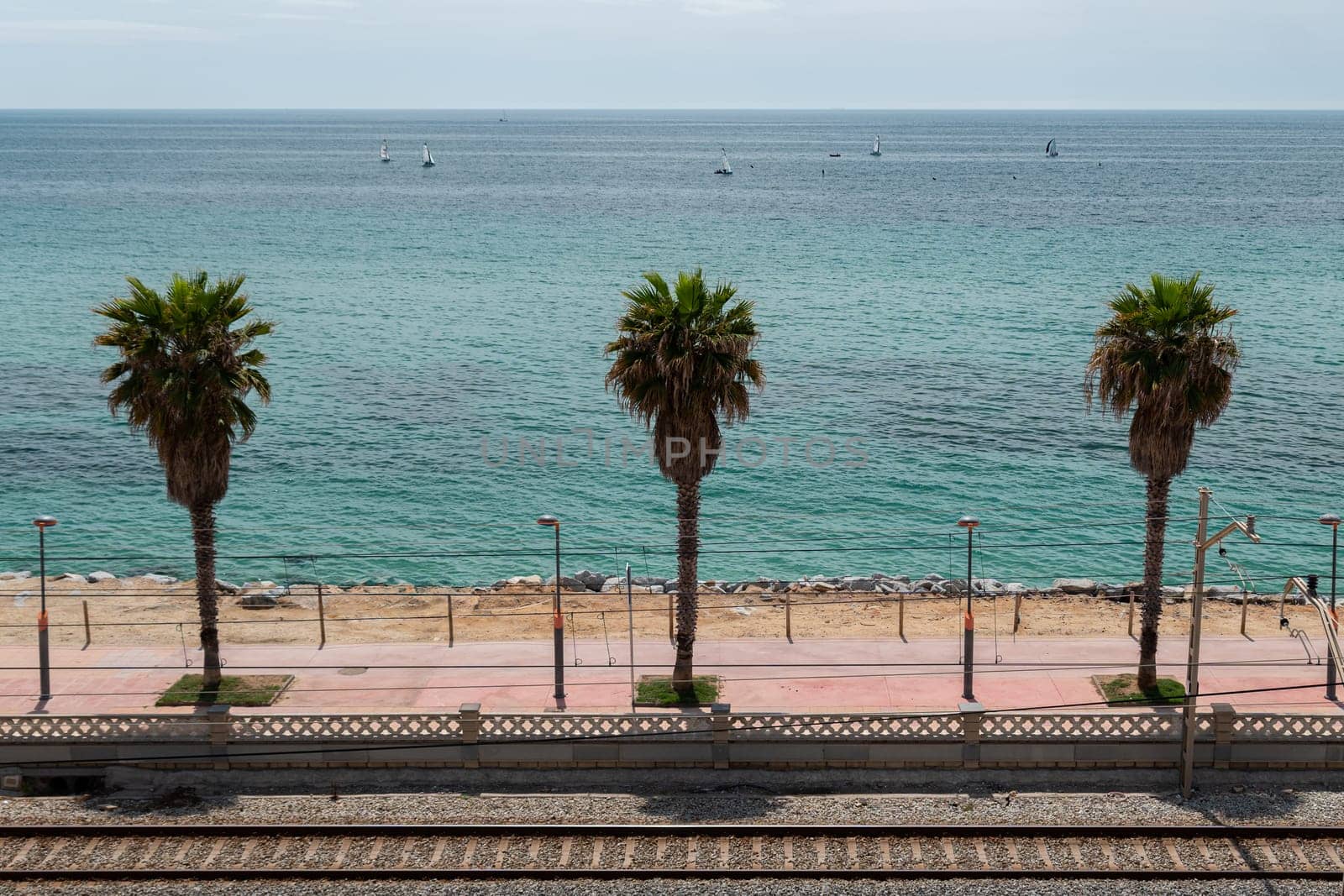 Tram railway and high palms on embankment in Vilassar de Mar, Barcelona on summer day. Picturesque cityscape with calm sea bay at Spanish resort