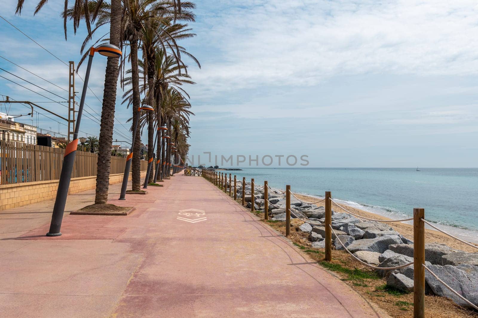 Waterfront with palms and street lamps row and sea sand beach with protective stones. Stylish villa buildings behind fence in Vilassar de Mar