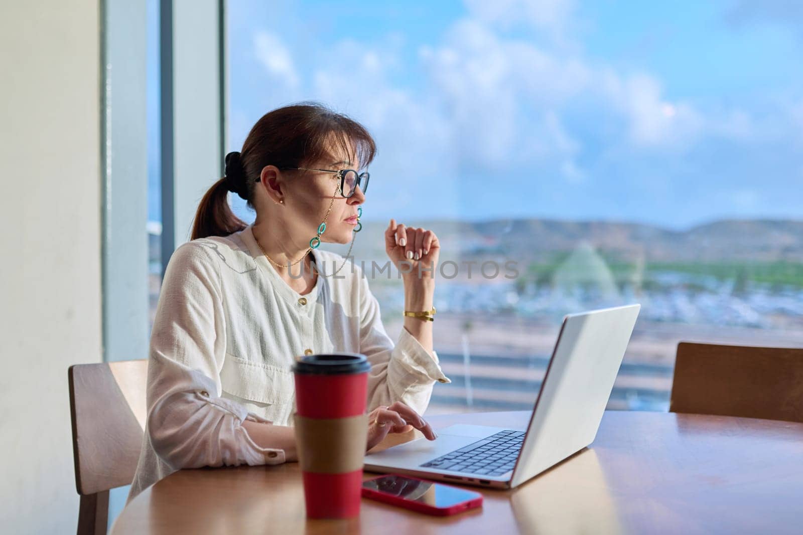 Middle-aged woman in an airport cafe working with a laptop computer, sitting at table with takeaway coffee, near panoramic window