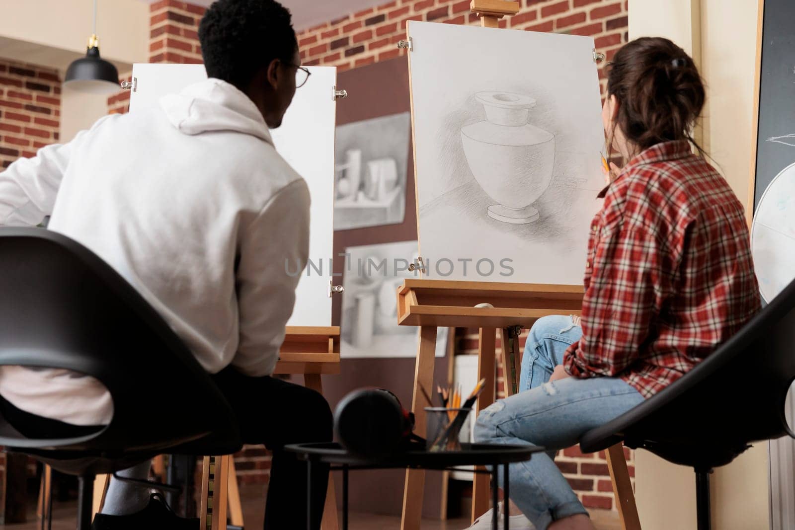 Young diverse people students looking at canvas with vase sketch, develop skills in line and shape while studying in art academy. Multiracial couple sitting at easels learning to draw together