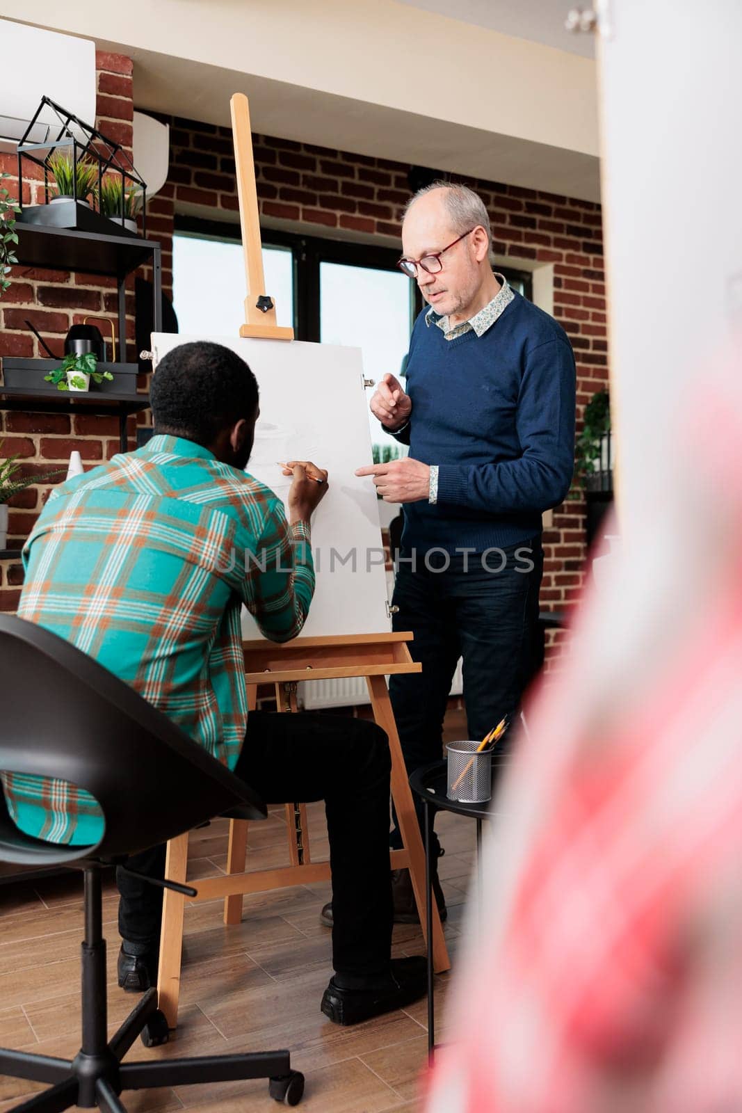 Senior man art teacher standing near easel working with African American male student during drawing lesson, experienced instructor teaching people to sketch. Artistic creative activities for adults