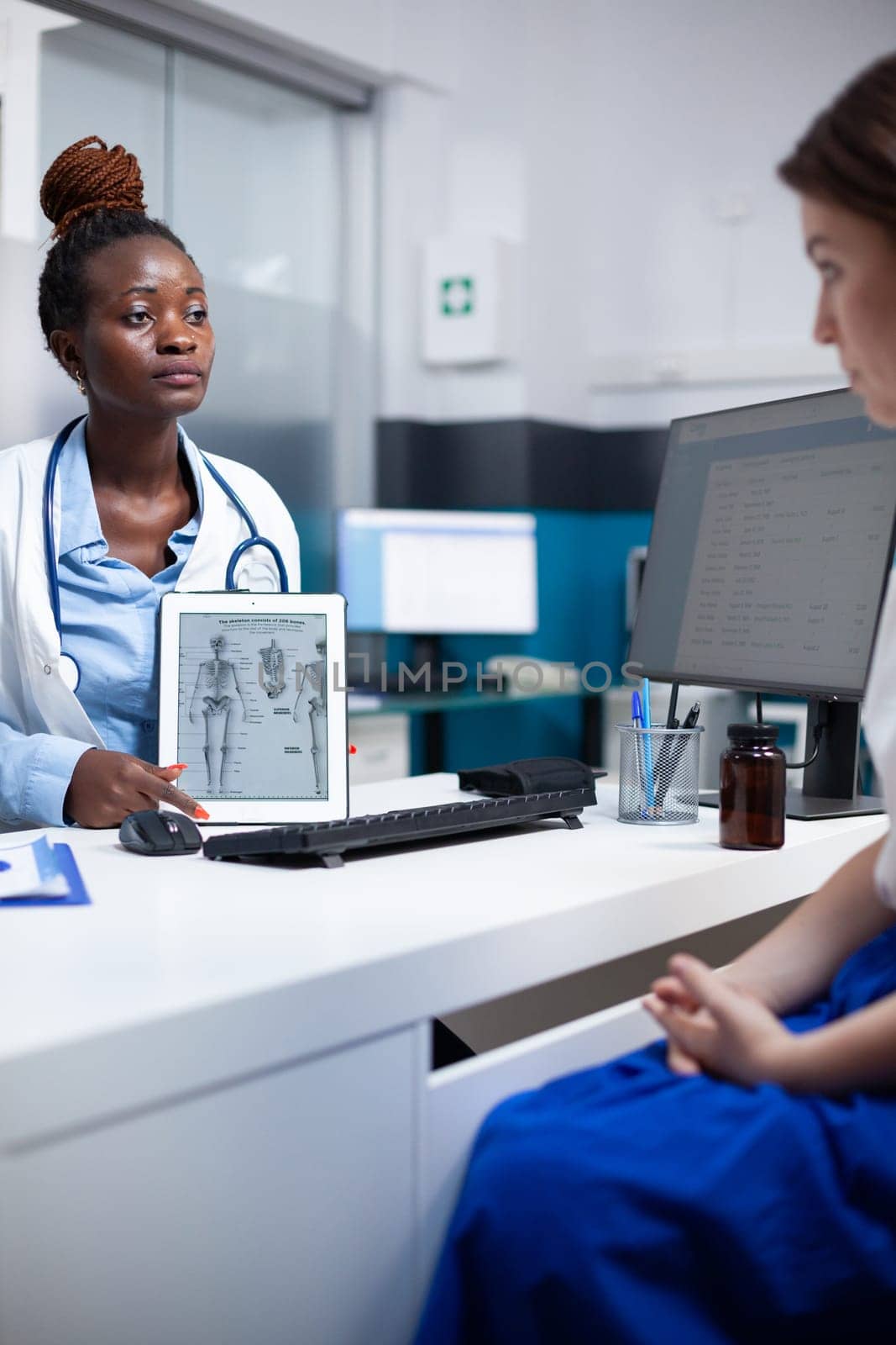 Bone specialist doctor at physical consultation offering anatomy information to patient on tablet using human skeleton figure. Orthopedist providing healthcare diagnosis to woman at appointment