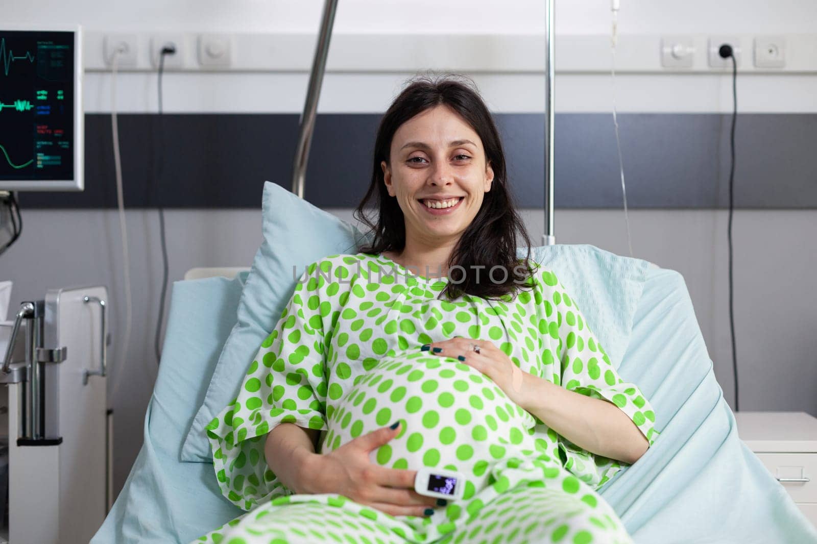 Smiling pregnant woman lying in bed holding hands on belly in hospital ward. Cheerful patient with belly pregnancy looking at camera and waiting on doctor to do examination during checkup visit