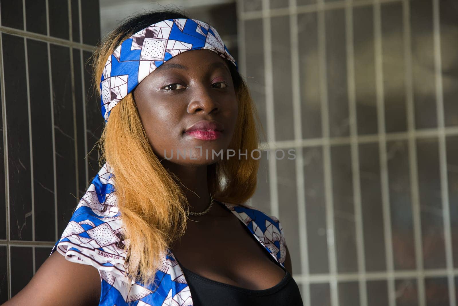 Outdoor portrait of a black girl wearing a smiling blue and white scarf standing against a building wall and looking at the camera.