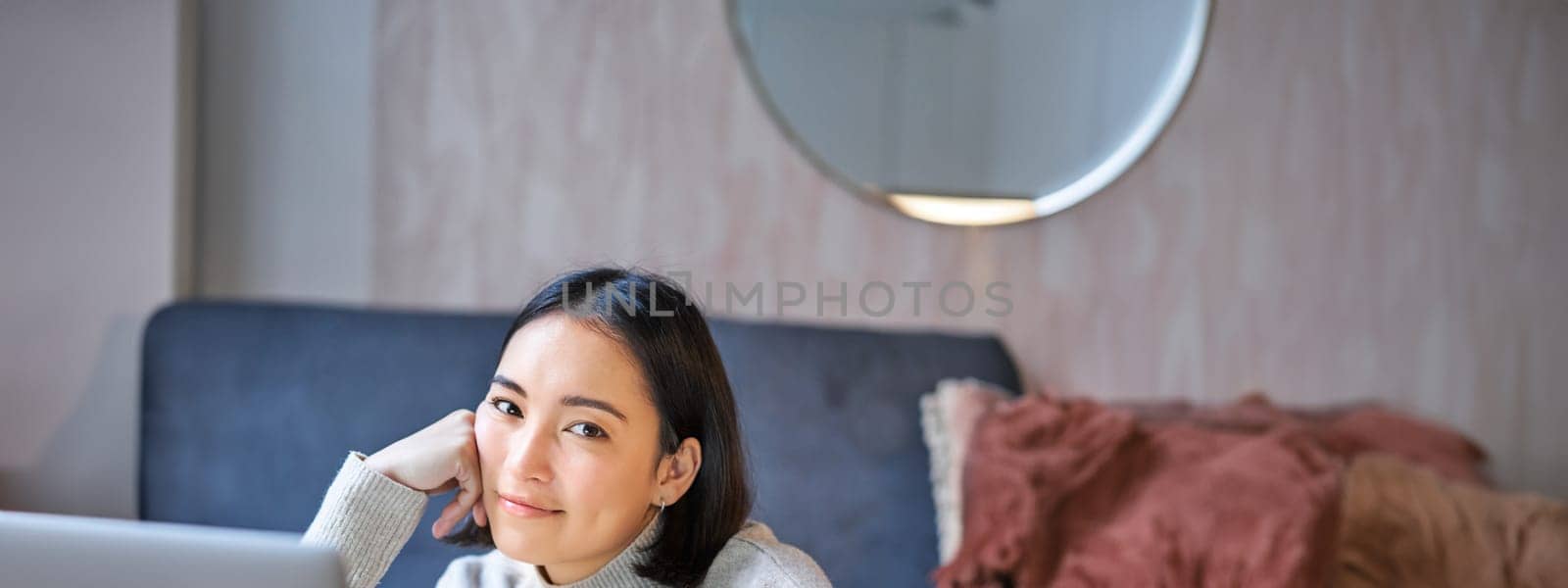 Vertical shot of young asian woman looking tired, smiling with exhausted expression, working from home on her laptop.