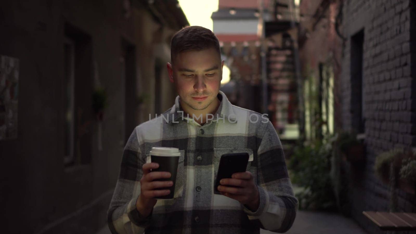 A young man walks along a narrow street late at night chatting on his smartphone and drinking coffee. A man in a warm shirt walks with a phone in his hands and drinks a hot drink. 4k