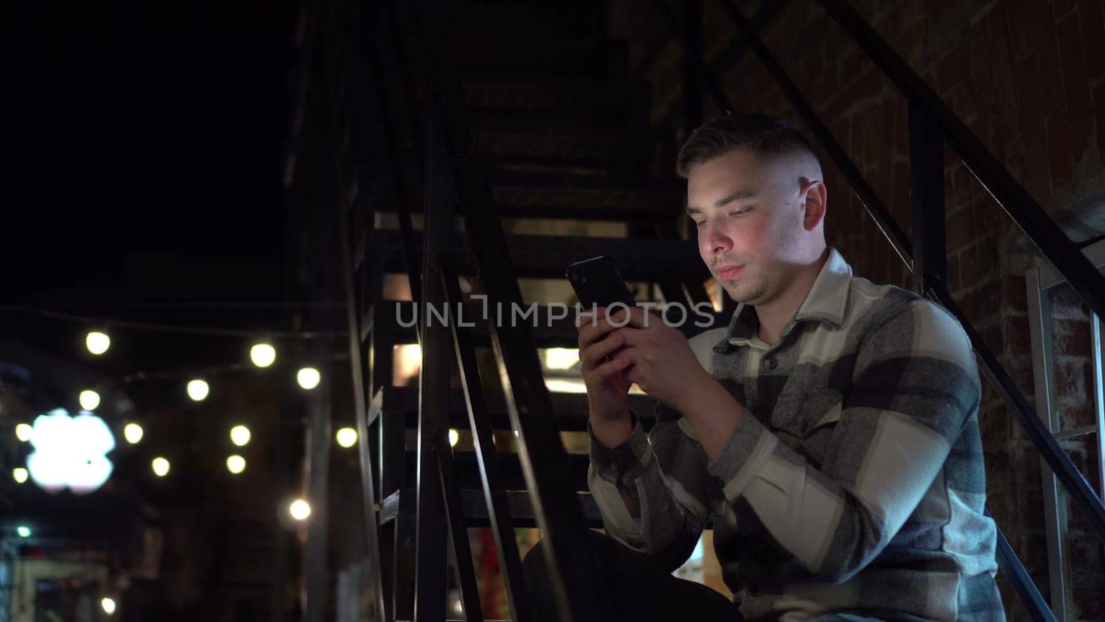 A young man sits on a fire escape and texts on a smartphone. A man on a narrow street late at night with a phone in his hand. 4k