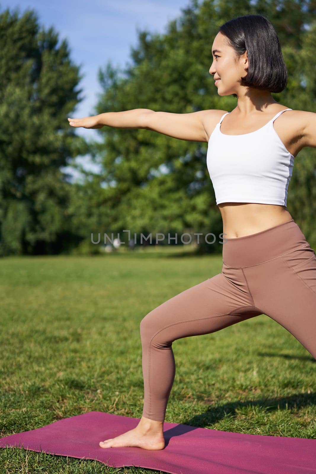 Young woman stretching her body, workout and doing yoga in park, standing on rubber mat in fitness clothing during training session by Benzoix