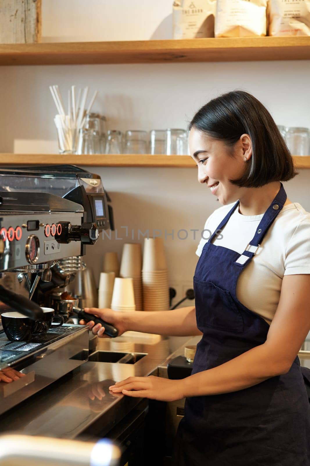 Happy beautiful asian girl, barista in apron works with coffee machine behind counter, standing in cafe and making cappuccino by Benzoix