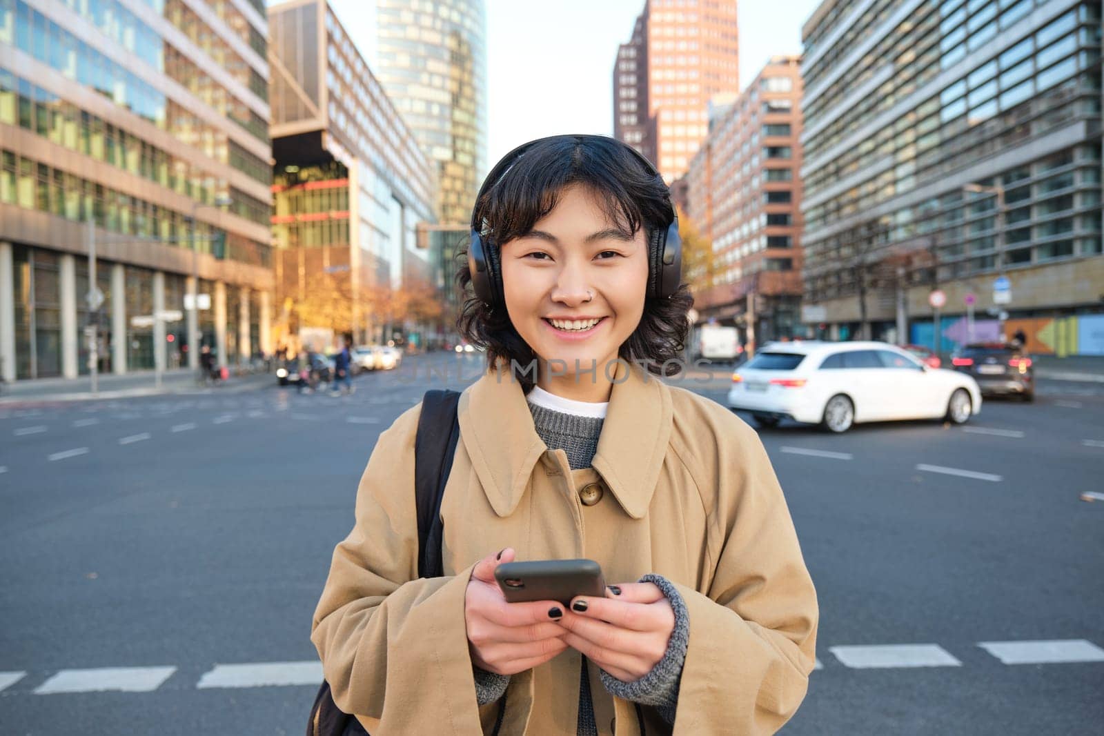 Portrait of young asian girl, student walks in city, listens music in headphones and uses mobile phone on streets.