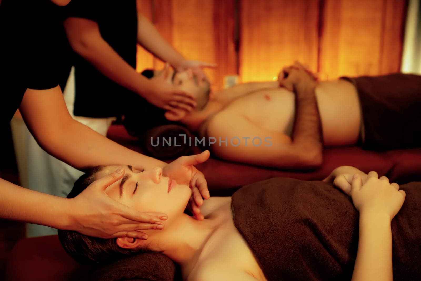 Couple customer enjoying relaxing anti-stress head massage and pampering facial beauty skin recreation leisure in warm candle lighting ambient salon spa in luxury resort or hotel. Quiescent