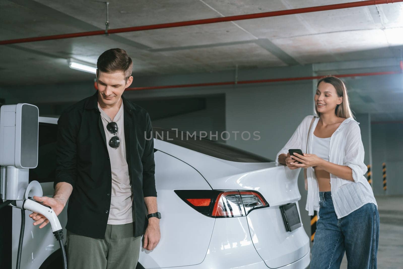 Young couple travel with EV electric car to shopping center parking lot charging in downtown city showing urban sustainability lifestyle by green clean rechargeable energy of electric vehicle innards