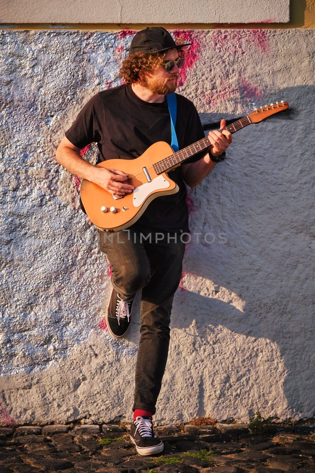 Hipster street musician in black playing electric guitar in the street on sunset leaning on a wall
