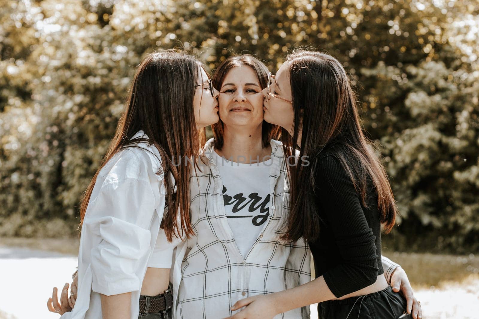 Portrait of a young Caucasian mother with two adult daughters who are kissing her on both cheeks while standing in a city park on a summer day, side view.