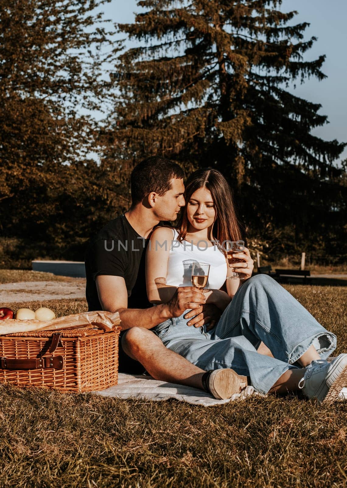 One beautiful caucasian young couple holding glasses of champagne sit hugging on a bedspread with a wicker basket and fruits in the park on a summer sunny day, close-up side view.