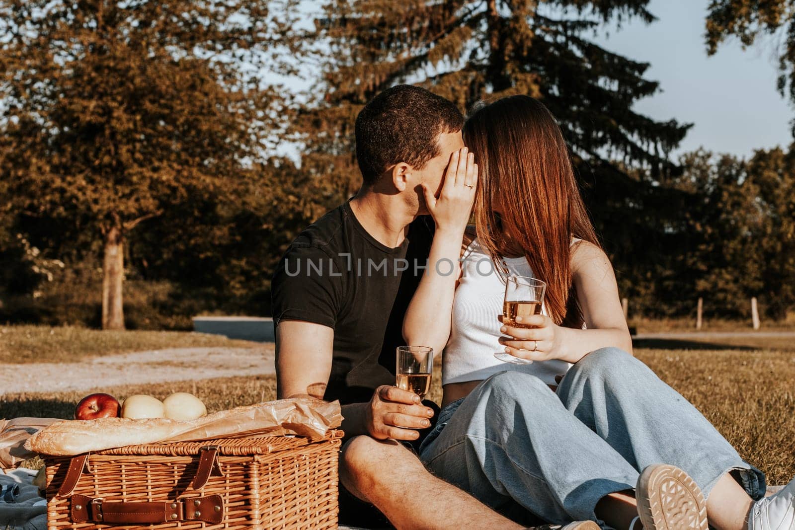 One beautiful caucasian young couple kissing covering their faces with their hands and holding glasses of champagne sit on a bedspread with a wicker basket and fruits in the park on a summer sunny day, close-up side view.