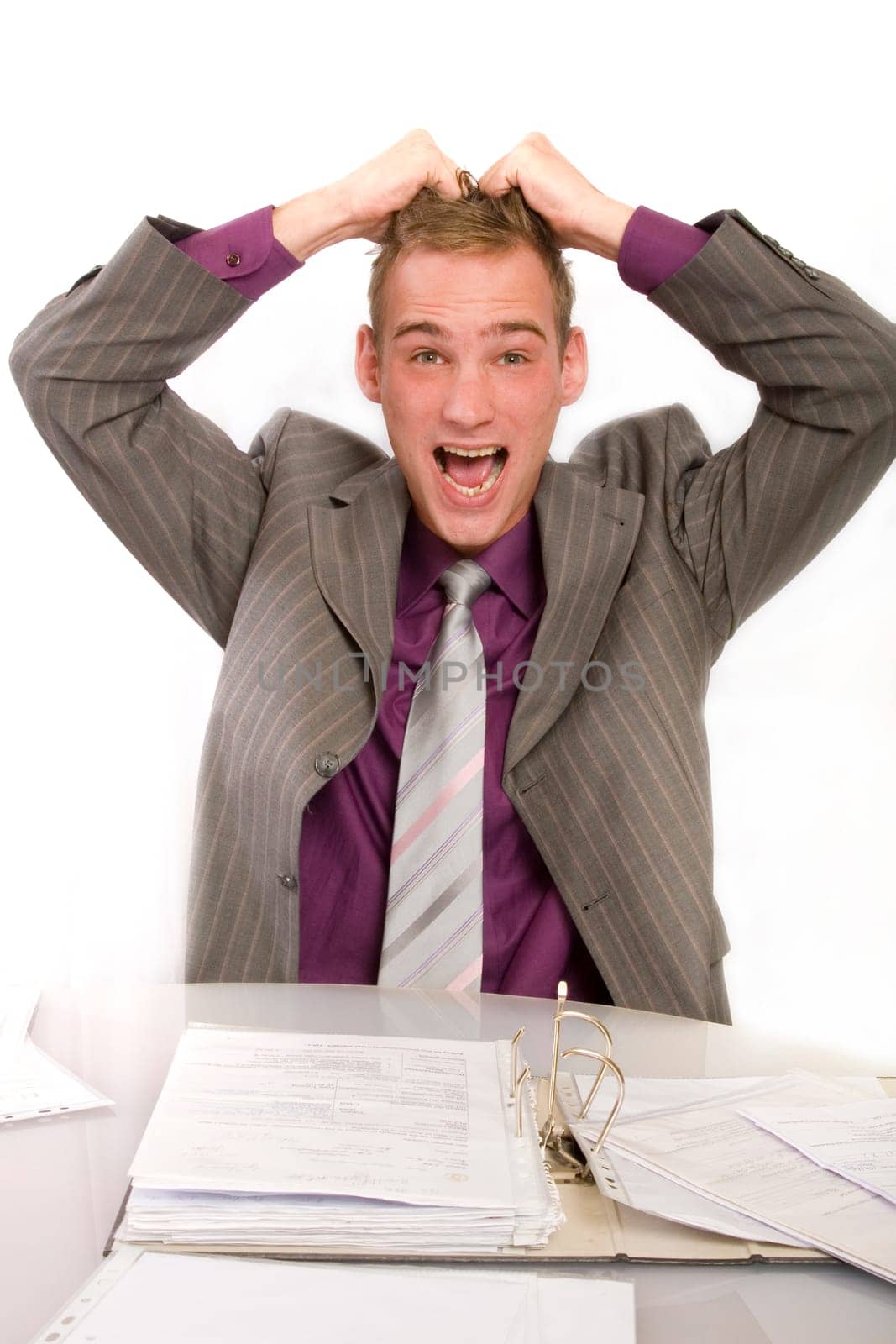 Desperate businessman in office at desk. He holds his hands above his head and screams. In front of him is a file folder.