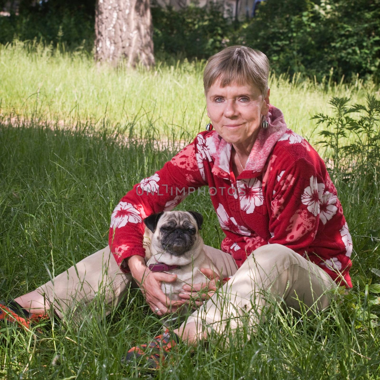 Full length view of elderly woman with dog (pug) and looking at camera sitting in grass in summer