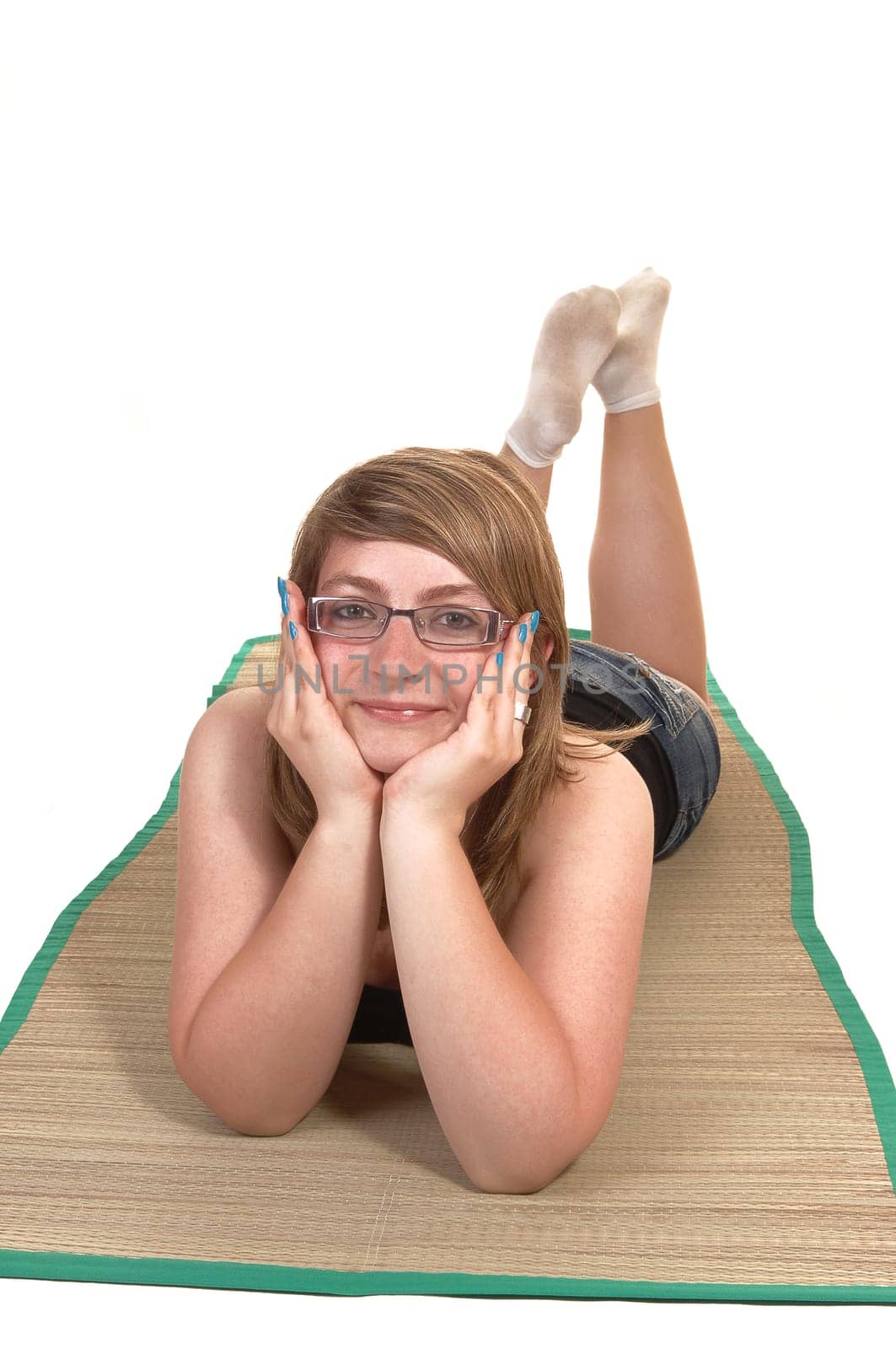 An relaxing young teenager lying on her stomach on a yoga matt on the  floor, her head in her hands and smiling into the camera, over white.