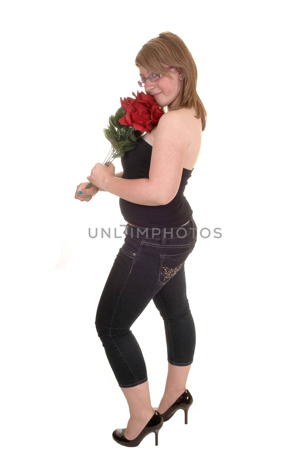 A pretty teenager standing in profile for white background, holding a bunch of red roses in her hand, in jeans and high heels.
